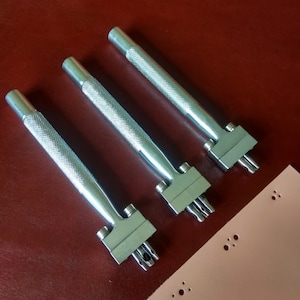 May include: Three silver leather hole punch tools with a textured handle. The tools are arranged in a row on a red surface. A piece of light pink leather with multiple rows of punched holes is in the lower right corner of the image.