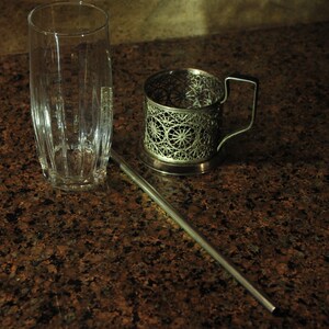 Puede incluir: Un portavasos de metal plateado con un dise&ntilde;o decorativo se encuentra sobre una encimera de granito marr&oacute;n. Un vaso transparente y una pajita de metal plateado tambi&eacute;n est&aacute;n sobre la encimera.