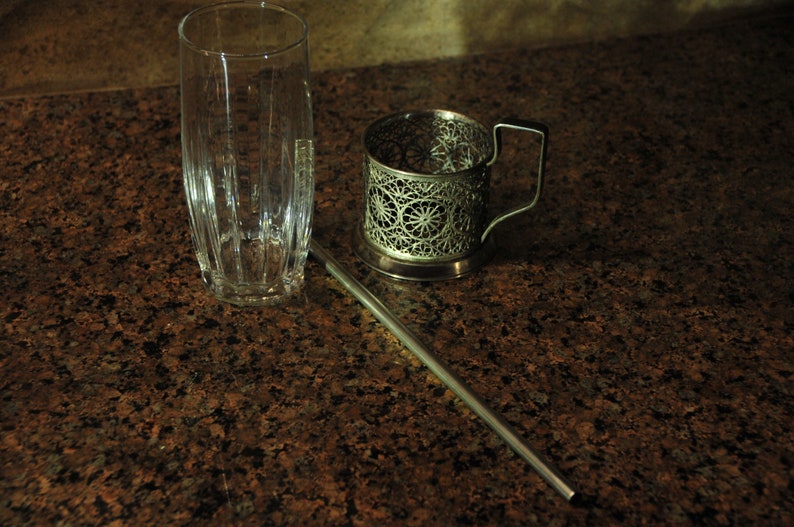 Puede incluir: Un portavasos de metal plateado con un dise&ntilde;o decorativo se encuentra sobre una encimera de granito marr&oacute;n. Un vaso transparente y una pajita de metal plateado tambi&eacute;n est&aacute;n sobre la encimera.