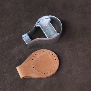 May include: A metal leather craft tool for cutting out teardrop shapes, with a matching teardrop shape cut out of brown leather.
