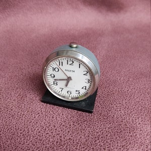 May include: A silver alarm clock with a white face and black numbers. The clock is labeled "PAKETA" and has a silver knob on the top.
