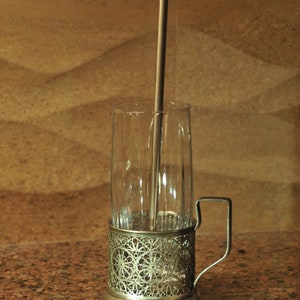 Puede incluir: Un portavasos de plata con un dise&ntilde;o decorativo y un asa. Una pajita de plata est&aacute; dentro de un vaso transparente.