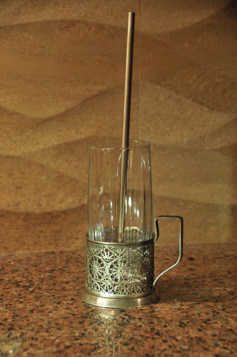 Puede incluir: Un portavasos de plata con un dise&ntilde;o decorativo y un asa. Una pajita de plata est&aacute; dentro de un vaso transparente.