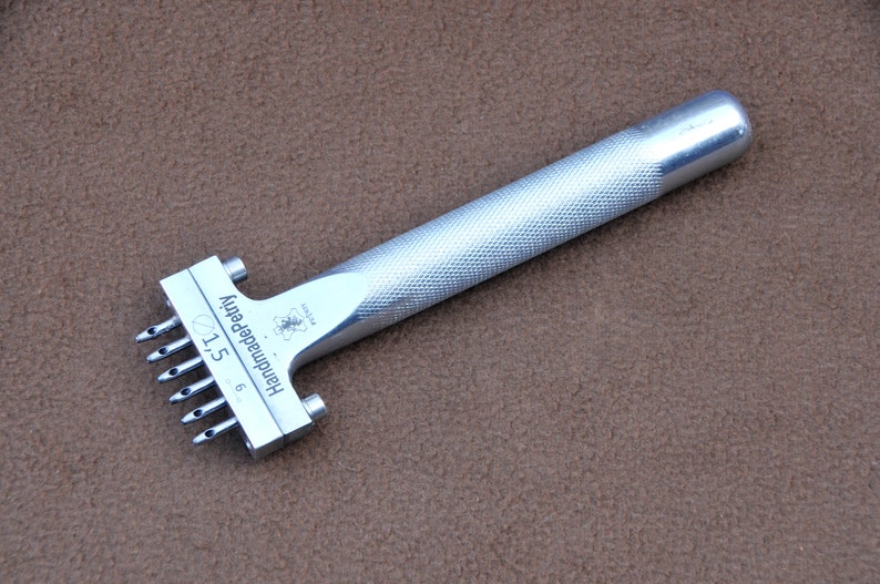 May include: Silver leather punch tool with a textured handle. The tool has six needles and is labelled "Handmade Petri 6/1.5".