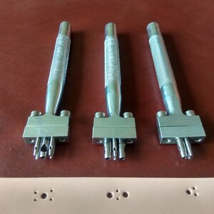 May include: Three silver leather hole punch tools with different sized punches. The tools are on a brown surface.