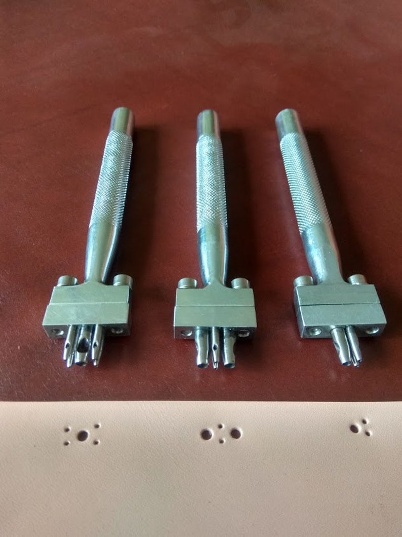 May include: Three silver leather hole punch tools with different sized punches. The tools are on a brown surface.