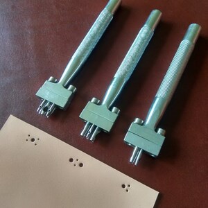 May include: Three silver leather hole punch tools with handles and multiple punch holes. A piece of light brown leather with multiple holes punched in it.
