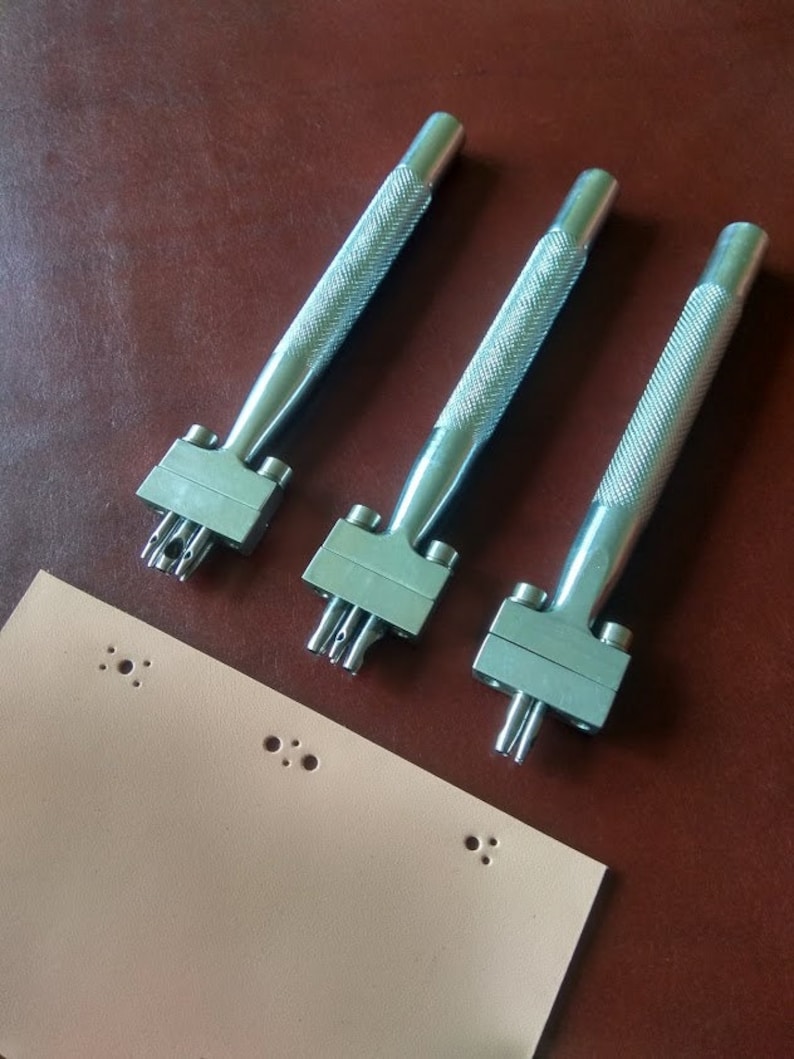 May include: Three silver leather hole punch tools with handles and multiple punch holes. A piece of light brown leather with multiple holes punched in it.