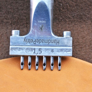 May include: A silver leather punch tool with the text "Handmade quality" and "1,5" printed on it. The tool has six prongs and is being used to punch holes in a piece of brown leather.
