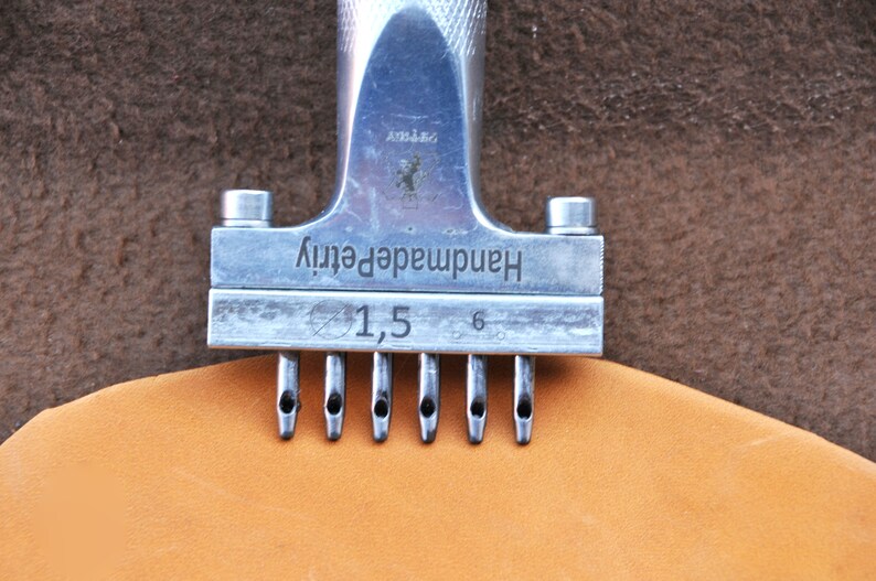 May include: A silver leather punch tool with the text "Handmade quality" and "1,5" printed on it. The tool has six prongs and is being used to punch holes in a piece of brown leather.