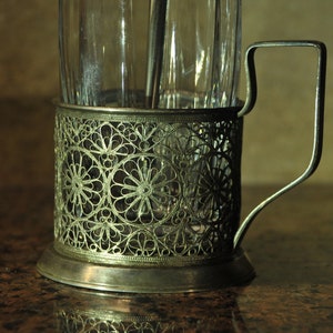 Puede incluir: Un portavasos de t&eacute; de metal plateado con un intrincado dise&ntilde;o floral. El portavasos tiene un asa y una base redonda.