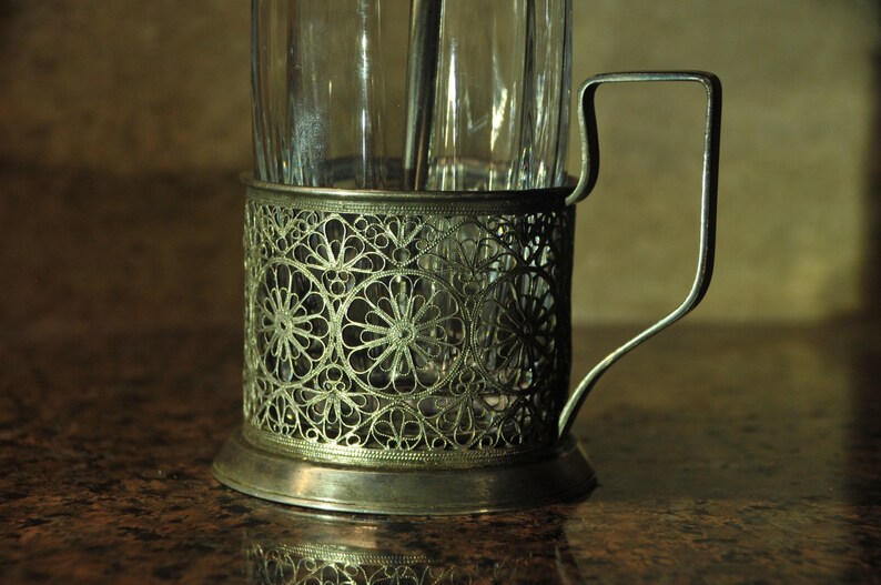 Puede incluir: Un portavasos de t&eacute; de metal plateado con un intrincado dise&ntilde;o floral. El portavasos tiene un asa y una base redonda.