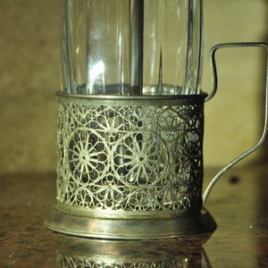 Puede incluir: Un portavasos de plata con un dise&ntilde;o delicado e intrincado. El portavasos tiene un asa y est&aacute; dise&ntilde;ado para sostener un vaso de t&eacute; o caf&eacute;.