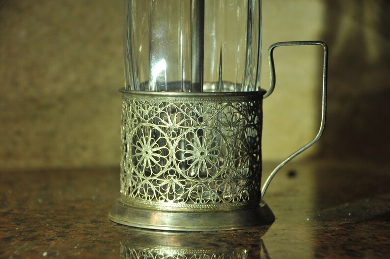 Puede incluir: Un portavasos de plata con un dise&ntilde;o delicado e intrincado. El portavasos tiene un asa y est&aacute; dise&ntilde;ado para sostener un vaso de t&eacute; o caf&eacute;.
