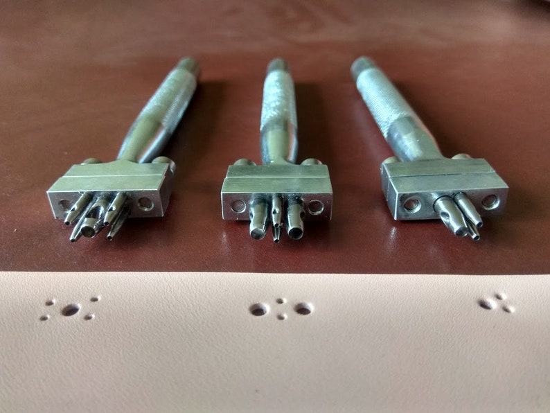 May include: Three silver leather hole punch tools with different sized punches. The tools are on a brown leather surface. The tools are used to create holes in leather for stitching or other purposes.