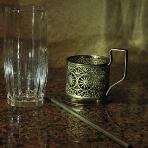 Puede incluir: Un portavasos de metal plateado con un dise&ntilde;o intrincado se encuentra sobre una encimera de granito marr&oacute;n junto a un vaso transparente. Una pajita de metal plateado se encuentra sobre la encimera.