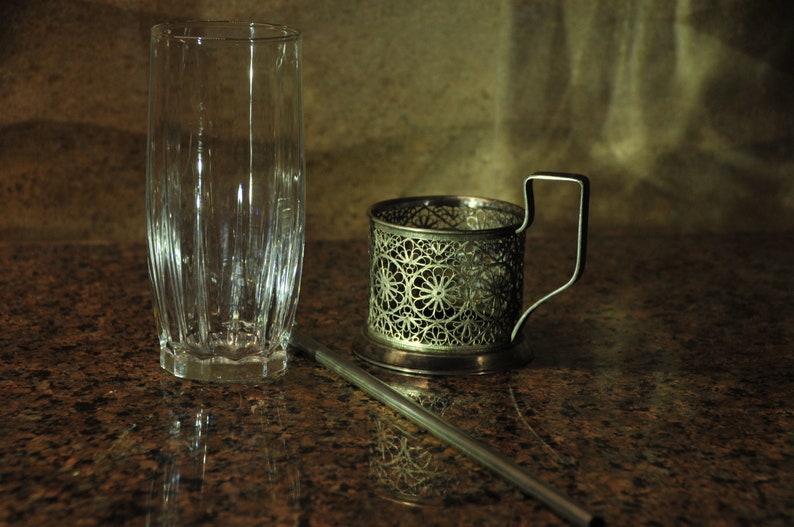 Puede incluir: Un portavasos de metal plateado con un dise&ntilde;o intrincado se encuentra sobre una encimera de granito marr&oacute;n junto a un vaso transparente. Una pajita de metal plateado se encuentra sobre la encimera.