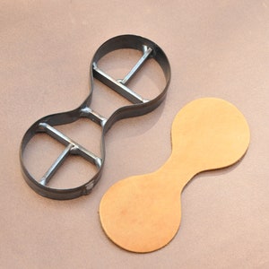 May include: A metal leather craft punch tool with a double-ended, figure-eight shape. The tool is next to a piece of brown leather cut in the same shape.