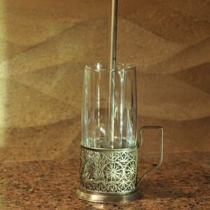 Puede incluir: Un portavasos de vidrio plateado con un dise&ntilde;o delicado e intrincado. El portavasos tiene un asa y un vaso de vidrio en su interior. Una pajita de metal est&aacute; dentro del vaso.