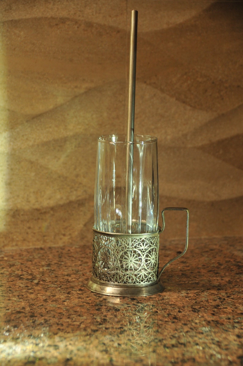 Puede incluir: Un portavasos de vidrio plateado con un dise&ntilde;o delicado e intrincado. El portavasos tiene un asa y un vaso de vidrio en su interior. Una pajita de metal est&aacute; dentro del vaso.