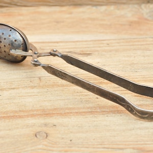 Puede incluir: Infusor de té de plata con un colador y un mango. El mango tiene un diseño decorativo.