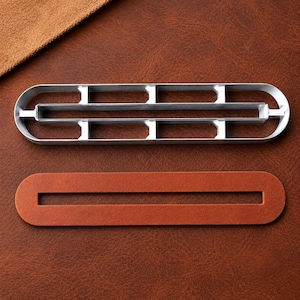 Peut inclure: Une matrice de découpe en métal argenté de forme ovale allongée avec une structure interne en grille, placée au-dessus d'une pièce en cuir marron assortie présentant une longue découpe rectangulaire étroite au centre de sa forme oblongue.