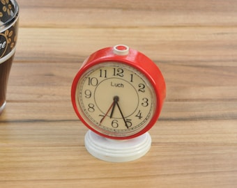 May include: A red alarm clock with a white face and black hands. The clock has a white base and the word "Luch" printed on the face.
