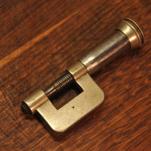 May include: A vintage brass and steel clamp with a threaded screw and a smooth, rounded handle. The clamp is designed to hold objects securely in place.