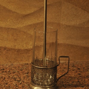 Puede incluir: Un portavasos de vidrio plateado con un dise&ntilde;o decorativo e intrincado. El portavasos tiene un asa y un cilindro de vidrio transparente en el interior. Una varilla de metal se extiende desde la parte inferior del portavasos hasta la parte superior del cilindro.