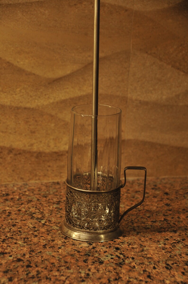 Puede incluir: Un portavasos de vidrio plateado con un dise&ntilde;o decorativo e intrincado. El portavasos tiene un asa y un cilindro de vidrio transparente en el interior. Una varilla de metal se extiende desde la parte inferior del portavasos hasta la parte superior del cilindro.