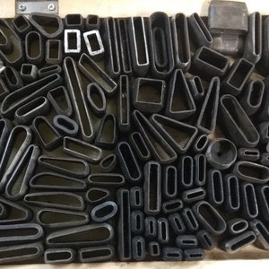 May include: A collection of metal shapes, including circles, ovals, squares, and rectangles, arranged in a grid pattern. The shapes are all black and have a rough, unfinished texture.