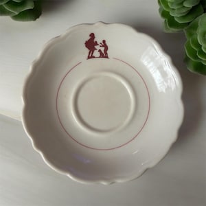 May include: White ceramic saucer with a scalloped edge and a red line around the center. A red silhouette of a woman and child with a dog is in the center.
