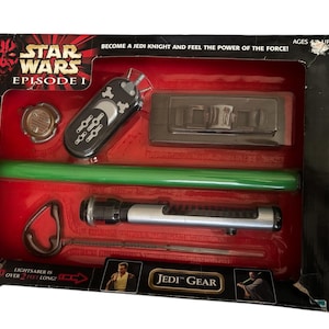 May include: A Star Wars Episode I Jedi Gear set with a lightsaber, a green lightsaber blade, a remote control, a small metal disc, and a black plastic device. The box is red and black with the words "Star Wars Episode I" and "Jedi Gear".