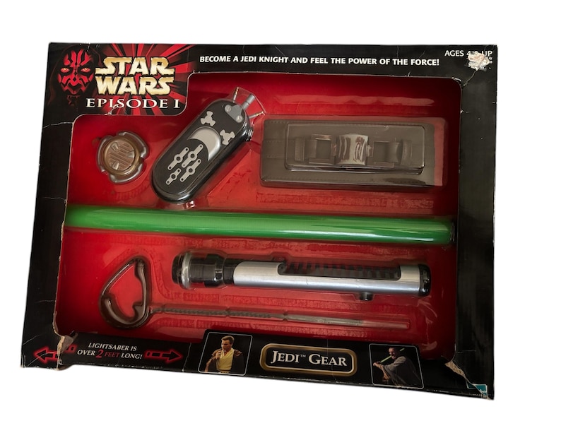 May include: A Star Wars Episode I Jedi Gear set with a lightsaber, a green lightsaber blade, a remote control, a small metal disc, and a black plastic device. The box is red and black with the words "Star Wars Episode I" and "Jedi Gear".