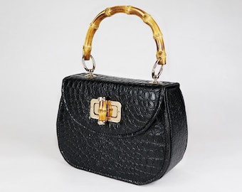Könnte beinhalten: Eine schwarze Handtasche aus krokodilgeprägtem Leder mit einem Bambus-Oberhenkel. Die Tasche hat einen goldfarbenen Verschluss und eine geschwungene Form.