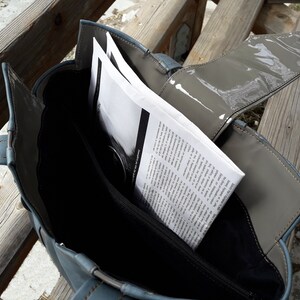 May include: A blue tote bag with a gray flap and a zipper closure. The bag is open and has a stack of papers inside.