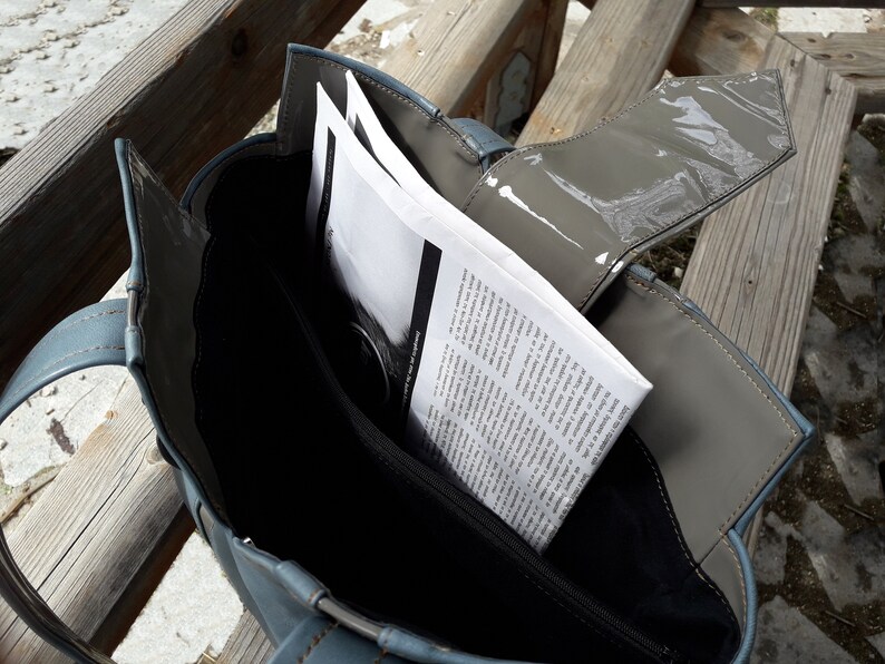 May include: A blue tote bag with a gray flap and a zipper closure. The bag is open and has a stack of papers inside.
