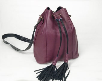 May include: A burgundy leather bucket bag with a black adjustable strap and black tassel accents.
