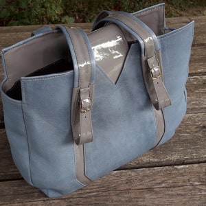 May include: A light blue tote bag with gray leather straps and a gray leather flap. The bag has a top zipper closure and a spacious interior.