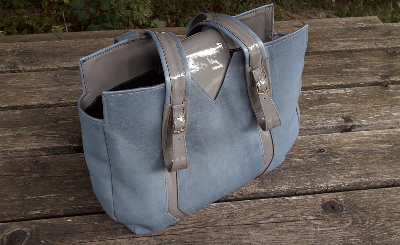 May include: A light blue tote bag with gray leather straps and a gray leather flap. The bag has a top zipper closure and a spacious interior.