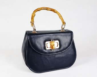 Könnte beinhalten: Marineblaue Handtasche mit Bambusgriff und goldfarbener Hardware. Die Tasche hat eine abgerundete Form und einen Klappenverschluss mit einem Bambus- und Goldverschluss. Der Griff ist gebogen.