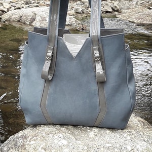 May include: A gray leather tote bag with gray leather straps and buckles. The bag is sitting on a rock near a stream.