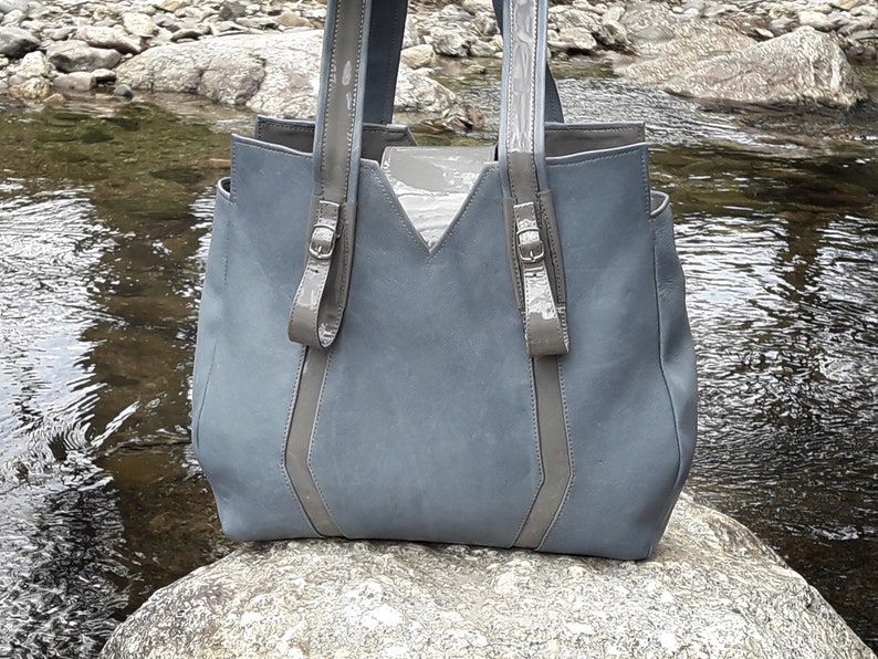 May include: A gray leather tote bag with gray leather straps and buckles. The bag is sitting on a rock near a stream.