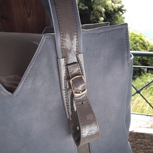 May include: A gray leather tote bag with a brown leather handle and a gold buckle. The bag has a V-shaped opening and a brown leather lining.