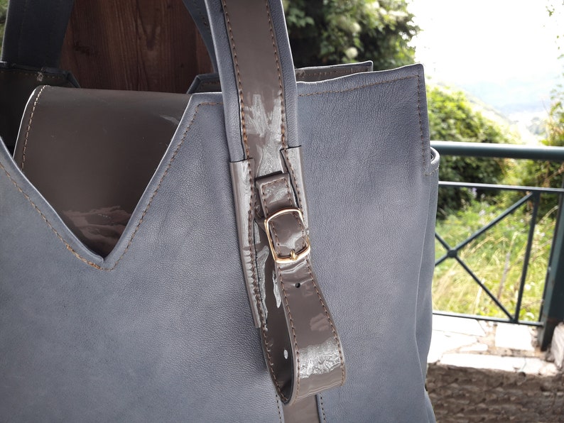 May include: A gray leather tote bag with a brown leather handle and a gold buckle. The bag has a V-shaped opening and a brown leather lining.