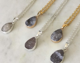 May include: A collection of teardrop-shaped pendant necklaces. The pendants feature a sparkling, crystal-like texture in shades of gray, white, and purple, set in silver and gold-toned metal. The necklaces are displayed on a white marble surface.