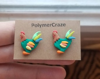 May include: A pair of polymer clay rooster earrings. The roosters are green with orange beaks and legs, and have colorful feathers. The earrings are on a brown card with the text "PolymerCraze".