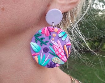 May include: Purple and pink statement earrings with a floral and abstract design. The earrings are made of polymer clay and have a round shape.
