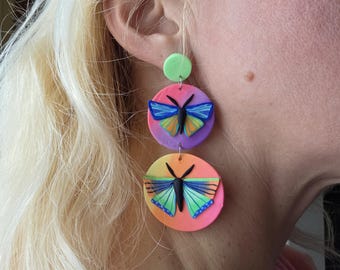May include: Colorful, handmade earrings featuring a layered design. Each earring has a green circle at the top, followed by two larger, round, gradient-colored discs. Each disc has a butterfly design with blue, orange, and black details. The earrings are a unique accessory.