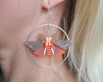 May include: A pair of hoop earrings with a red and brown moth charm. The moth has a detailed design with black and white accents.